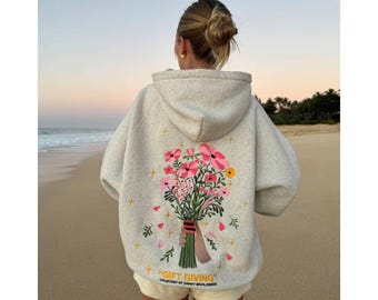 May include: A heathered beige hooded sweatshirt featuring a large graphic on the back of a hand holding a colorful bouquet of pink, red, and yellow flowers. The text "GIFT GIVING" is printed in yellow below the floral illustration.
