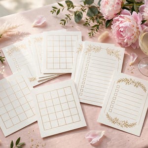 May include: A collection of white paper stationery cards featuring gold foil grid patterns and checklist designs with floral accents. The cards are arranged on a light pink surface alongside pink peony flowers and a glass of champagne.