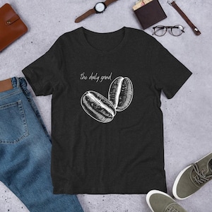 May include: Dark gray t-shirt featuring a white illustration of two coffee beans and the text "the daily grind". The shirt is made of a soft material and is suitable for casual wear. The shirt is displayed on a gray surface.