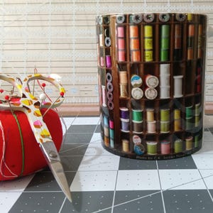 May include: A red tomato pin cushion with needles and pins, next to a mug with a sewing-themed design. The mug features images of spools of thread and sewing notions. A pair of decorative scissors rests on the table.