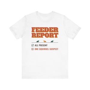 Op de afbeelding: Een wit t-shirt met korte mouwen met de tekst FEEDER REPORT in oranje, drie kleine vogelillustraties op een lijn en een checklist met de tekst ALL PRESENT en ONE SQUIRREL SUSPECT in donkergrijs en oranje.