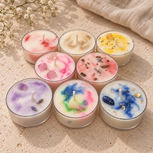 May include: A set of eight round tea light candles in clear plastic cups, each featuring unique wax swirls in red, yellow, pink, purple, blue, and green, decorated with embedded dried flower petals, small crystals, and gold leaf flakes.