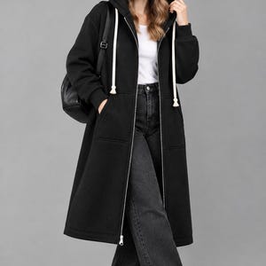 Könnte beinhalten: Ein langer schwarzer Kapuzenpullover mit Reißverschluss, Kapuze mit dicken weißen Kordelzügen, langen Ärmeln mit gerippten Bündchen und zwei Vordertaschen. Das Kleidungsstück wird über einem weißen Oberteil und einer dunkelgrauen weiten Jeans mit weißen Turnschuhen getragen.