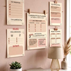 Könnte beinhalten: Ein Set aus fünf beigen Planer-Blättern aus Papier mit rosa Akzenten, die mit Klammern an einer Schnur hängen. Die Blätter enthalten Überschriften wie Top Priorities, Care & Ratitude Planner, Self-Reflection, Idea Seedling und Project Planning.