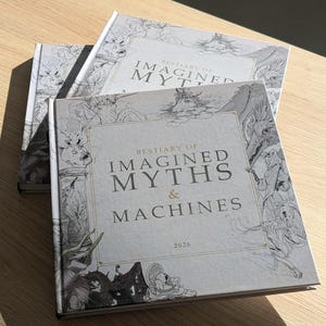 Pode incluir: Dois livros quadrados de capa dura com capas cinza-claras apresentando ilustrações complexas de linhas pretas de criaturas míticas e elementos mecânicos. O título diz Bestiary of Imagined Myths & Machines 2026 em fonte serifada.