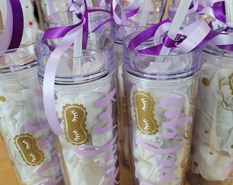 May include: Clear plastic tumblers with lids, straws, and purple ribbons. Each tumbler has a gold sleep mask design and the words "Bride Tribe" in purple. The tumblers are filled with white tissue paper.