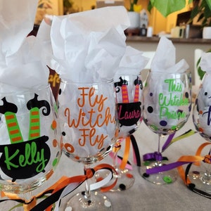 Könnte beinhalten: Klare Weingläser mit Halloween-Dekorationen. Jedes Glas hat ein einzigartiges Design, darunter Hexenhüte, Kessel und die Aufschriften "Fly Witch Fly" und "This Witch Poisons". Die Gläser sind mit bunten Bändern verziert.