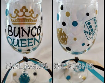 Puede incluir: Dos copas de vino transparentes con diseños decorativos. Una copa presenta una corona dorada y las palabras "Bunco Queen" en negro y turquesa. La otra copa tiene dados y lunares en dorado, turquesa y negro. Ambas copas tienen una cinta atada alrededor del tallo.