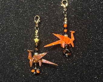 May include: Two orange paper crane charms featuring gold-tone lobster clasps. Each charm is accented with small beads in black, white, and orange, and one includes a small gold-tone star charm. The cranes are folded from iridescent paper.