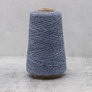 May include: A large conical spool of yarn featuring a heathered blue and white speckled texture. The yarn is wound tightly around a central cardboard core, displaying a consistent multi-tonal appearance throughout the entire length of the thread.
