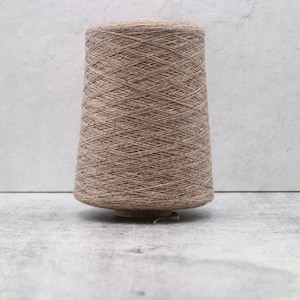 May include: A large, conical spool of light brown, textured yarn. The thread is wound tightly in a crisscross pattern around the central core, creating a uniform, dense appearance across the entire surface of the spool from top to bottom.