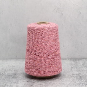 May include: A large conical spool of light pink yarn featuring a subtle speckled texture with tiny flecks of orange and white throughout the thread. The yarn is wound tightly around a central cardboard core, creating a uniform and dense structure.