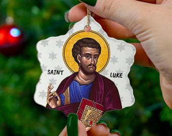 以下が含まれることがあります： 星型のクリスマスオーナメント。聖ルカのイメージと「SAINT」と「LUKE」の文字が描かれています。オーナメントは金の光輪があり、赤いオーナメントが付いたクリスマスツリーの前に飾られています。