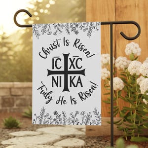 以下が含まれることがあります： 黒文字と花の縁取りのある白いガーデンの旗。テキストは「キリストは復活した！IC XC NIKA 真に彼は復活した！」