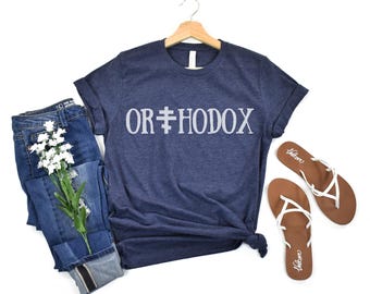以下が含まれることがあります： 白字で「ORTHODOX」とプリントされたネイビーブルーのTシャツ。 Tシャツは、ジーンズ、白と茶色のサンダル、白い花の小枝と一緒に表示されています。