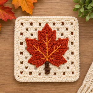 May include: A square crochet granny square coaster featuring an off-white yarn base with a central three-dimensional maple leaf motif in shades of burnt orange and red, accented by a dark brown stem and yellow embroidered leaf veins.
