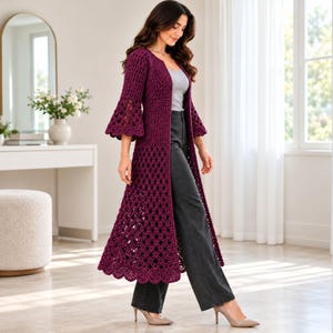 May include: A long, deep purple crochet duster cardigan featuring an open front, three-quarter length bell sleeves, and a detailed scalloped hemline. The garment is crafted with a textured stitch pattern that transitions into an intricate openwork design.