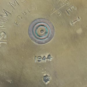 May include: A circular brass base of a 75mm M5A1 Type I artillery shell casing. The metal surface features concentric circular grooves and stamped text including 75MM M5A1-TYPEI, ECBC-1, and the year 1944. A central percussion primer is visible.