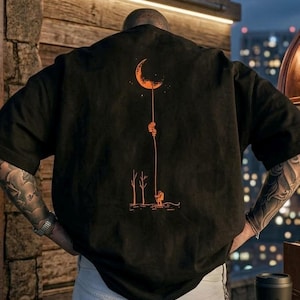 Puede incluir: Una camiseta negra de manga corta con un gráfico naranja en la espalda. El diseño muestra una luna creciente conectada por una línea larga a una pequeña figura junto a dos árboles desnudos, con formas circulares flotando arriba.