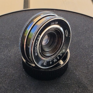 May include: A black Industar-69 camera lens with silver accents and a ridged focus ring. The front element displays the text 2.8/28 and the Cyrillic text ИНДУСТАР-69, along with aperture markings ranging from 2.8 to 16. The lens rests on a black cap.