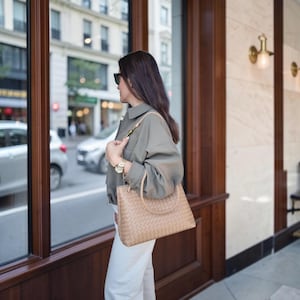 Può includere: Una borsa a mano in pelle intrecciata color beige con due manici arrotondati e una tracolla rimovibile. La borsa presenta una base rettangolare strutturata e una dettagliata trama intrecciata su tutta la superficie esterna, con finiture dorate.