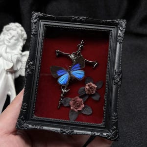 May include: A rectangular black frame with ornate detailing contains a deep red velvet backing. Inside, a metallic cross is adorned with a vibrant blue and black butterfly and two small, dark-toned artificial roses with leaves.