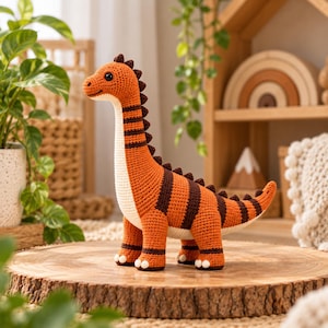 Könnte beinhalten: Ein gehäkeltes Dinosaurier-Spielzeug mit langem Hals und Schwanz, gestaltet mit horizontalen Streifen in Orange und Dunkelbraun. Die Figur hat einen cremefarbenen Bauch, eine Reihe kleiner dunkelbrauner Stacheln auf dem Rücken und zwei schwarze Perlenaugen.