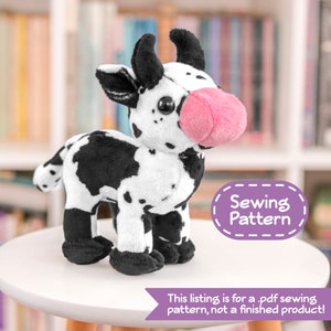 Könnte beinhalten: Ein schwarzes und weißes Plüsch-Kuh-Spielzeug mit einer rosa Nase. Das Spielzeug steht auf einem weißen Tisch. Der Text "Sewing Pattern" befindet sich auf einem lilafarbenen Etikett im Hintergrund.
