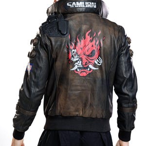 Puede incluir: Una chaqueta de cuero negra y marrón con un gran gráfico rojo y blanco de una máscara de demonio estilizada en la espalda. El cuello muestra el texto SAMURAI en letras blancas. La chaqueta incluye detalles negros en los hombros.