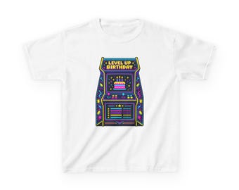 May include: A white t-shirt featuring a graphic of a retro arcade cabinet in neon colors. The screen displays a birthday cake with candles and the text LEVEL UP BIRTHDAY. The cabinet is decorated with stars, geometric shapes, and buttons.
