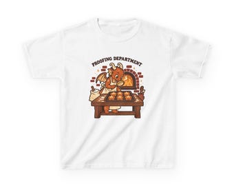 May include: A white t-shirt featuring a graphic of a small orange dragon wearing an apron, standing at a wooden table with a tray of star-shaped cookies. The text PROOFING DEPARTMENT is printed above the dragon, with a brick oven behind.