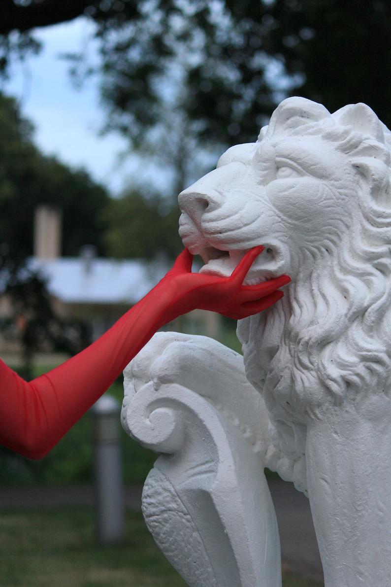 May include: A white plaster lion statue with a red glove touching its mouth. The statue is in a garden setting.