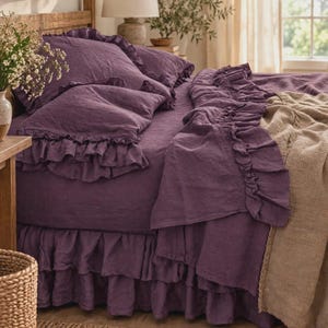 May include: A deep purple bedding set featuring a ruffled duvet cover, matching ruffled pillowcases, and a ruffled bed skirt. The fabric has a visible linen texture. A textured beige throw blanket is draped over the side of the bed.