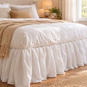 Può includere: Una balza per letto bianca con un design a volant arricciato in tessuto. La balza è posizionata sotto un copripiumino bianco, una coperta beige e diversi cuscini nelle tonalità del bianco e del marrone chiaro su una struttura letto.