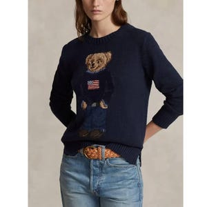 Op de afbeelding: Een marineblauwe trui met lange mouwen en ronde hals, voorzien van een centraal intarsia-gebreid teddybeer-motief. De beer draagt een donkere trui met een kleine Amerikaanse vlag en een blauwe broek. De trui heeft geribbelde boorden en zoom.