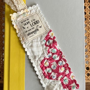 May include: A rectangular fabric bookmark featuring a cream-colored patch with the text 'but those who HOPE in the LORD will renew their strength (ISAIAH 40:31)'. The bookmark has a scalloped white edge, a cream lace ribbon, and a pink patterned section.