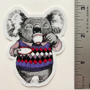 Puede incluir: Una ilustración en blanco y negro de un koala que lleva un suéter morado con un patrón de rombos rojos y blancos, sosteniendo una taza de té roja y blanca y un platillo. El koala tiene una expresión relajada y disfruta de una taza de té.