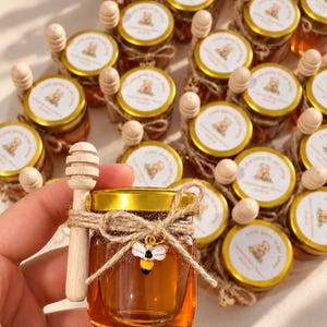 Puede incluir: Un pequeño frasco de vidrio lleno de miel ámbar, con tapa de metal dorado y etiqueta circular blanca con ilustración de oso y texto. Un cucharón de madera para miel está atado al frasco con cordel y un pequeño dije de abeja esmaltado.