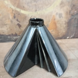 Puede incluir: Un jarrón de cerámica negra brillante con una forma cónica y geométrica que presenta pliegues angulares nítidos y una abertura circular en la parte superior. La superficie tiene un acabado reflectante que resalta el diseño estructurado y facetado.