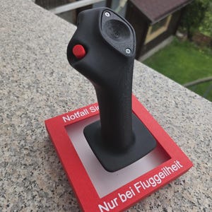 May include: A black joystick controller mounted on a square red base. The base features white text reading Notfall Si and Nur bei Fluggeilheit. The joystick has a textured matte finish, a small red button on the side, and a circular top panel secured with two silver screws.
