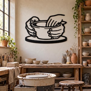 May include: A black metal wall art piece featuring a stylized line drawing of two hands shaping a clay bowl on a pottery wheel. The design is minimalist, showcasing the fingers and palms molding the circular vessel on a flat base.