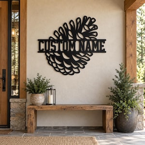 May include: A black metal wall sign shaped like a pinecone with a horizontal bar across the center featuring the text CUSTOM NAME in a bold, block font. The sign is mounted on a light-colored wall above a wooden bench.