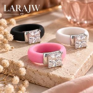 May include: Three silicone rings with square-cut clear stones set in silver-toned metal. The bands are black, white, and pink. The text LARAJW is visible at the top left of the image. The rings are arranged on a textured stone surface.
