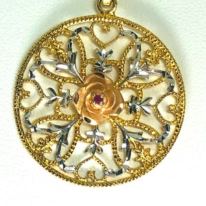 May include: A gold pendant with a filigree design featuring a rose with a red gemstone in the center. The pendant is made of two-toned gold, with a white gold filigree design and a yellow gold rose.