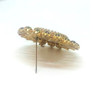 May include: A gold-toned brooch with a large, round, white center surrounded by a ring of green and black rhinestones. The brooch has a pin back.