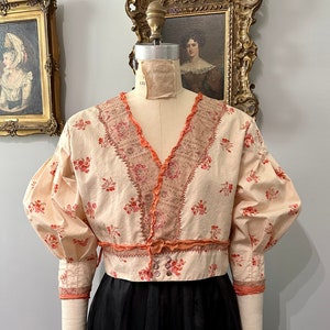 May include: A vintage-style blouse with a peach-colored background and a floral print. The blouse has a V-neckline, puffed sleeves, and a lace trim. The blouse is cinched at the waist with a ribbon.