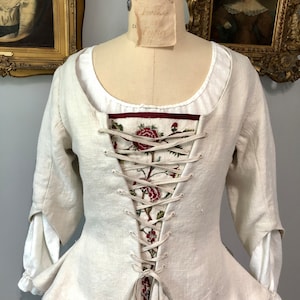 May include: A white linen bodice with a fitted waist and long sleeves. The bodice has a decorative floral embroidery panel in the front and is laced up the back with white ribbon.