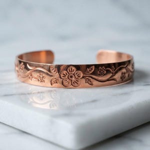 May include: A copper cuff bracelet featuring an embossed floral pattern with vines and leaves. The metal has a polished finish and an open back design, showcasing a repeating motif of small flowers centered around a larger bloom.