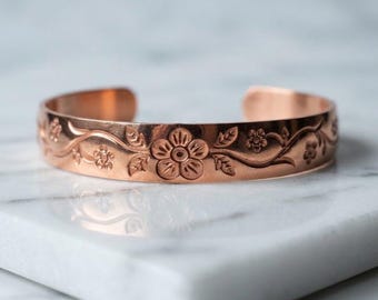 May include: A copper cuff bracelet featuring an embossed floral pattern with vines and leaves. The metal has a polished finish and an open back design, showcasing a repeating motif of small flowers centered around a larger bloom.
