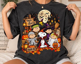 May include: A charcoal gray t-shirt featuring a colorful Halloween graphic with Peanuts characters dressed in costumes, a haunted house, a full moon, pumpkins, and the text "TRICK OR TREAT" and "BOO" surrounded by autumn leaves.
